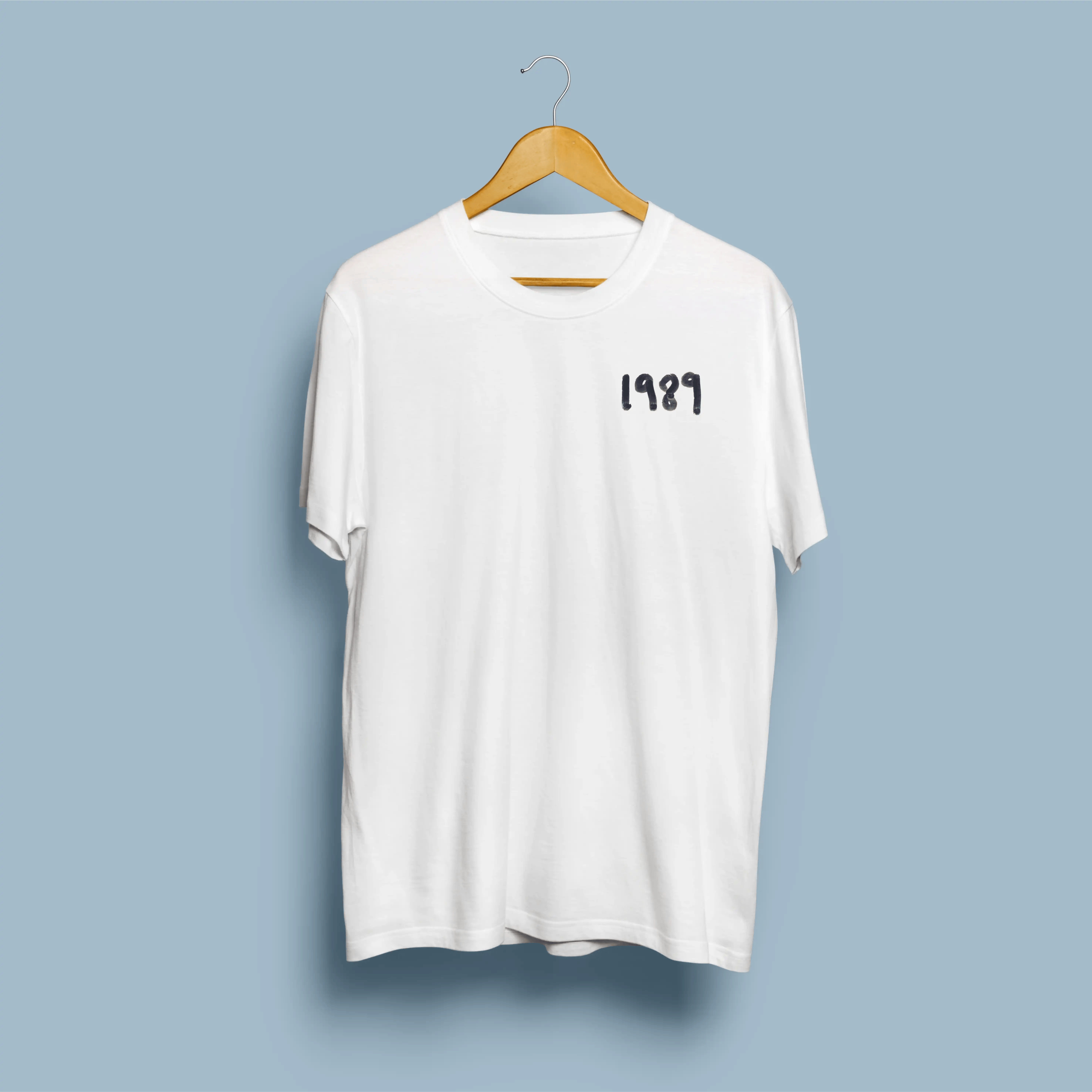 T-Shirt Taylor Swift - 1989 2 Frente de t-shirt branca com tipografia 1989 em graffiti azul-escuro no peito esquerdo, em cabide