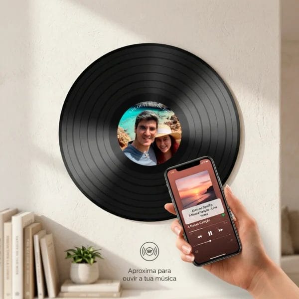 Disco de vinil personalizado com foto de casal, texto 'Nossa Música' e data '6 DE OUTUBRO DE 2021', pendurado na parede. Uma mão aproxima um smartphone para ativar a tag NFC escondida e ouvir música no Spotify via tag NFC invisível.